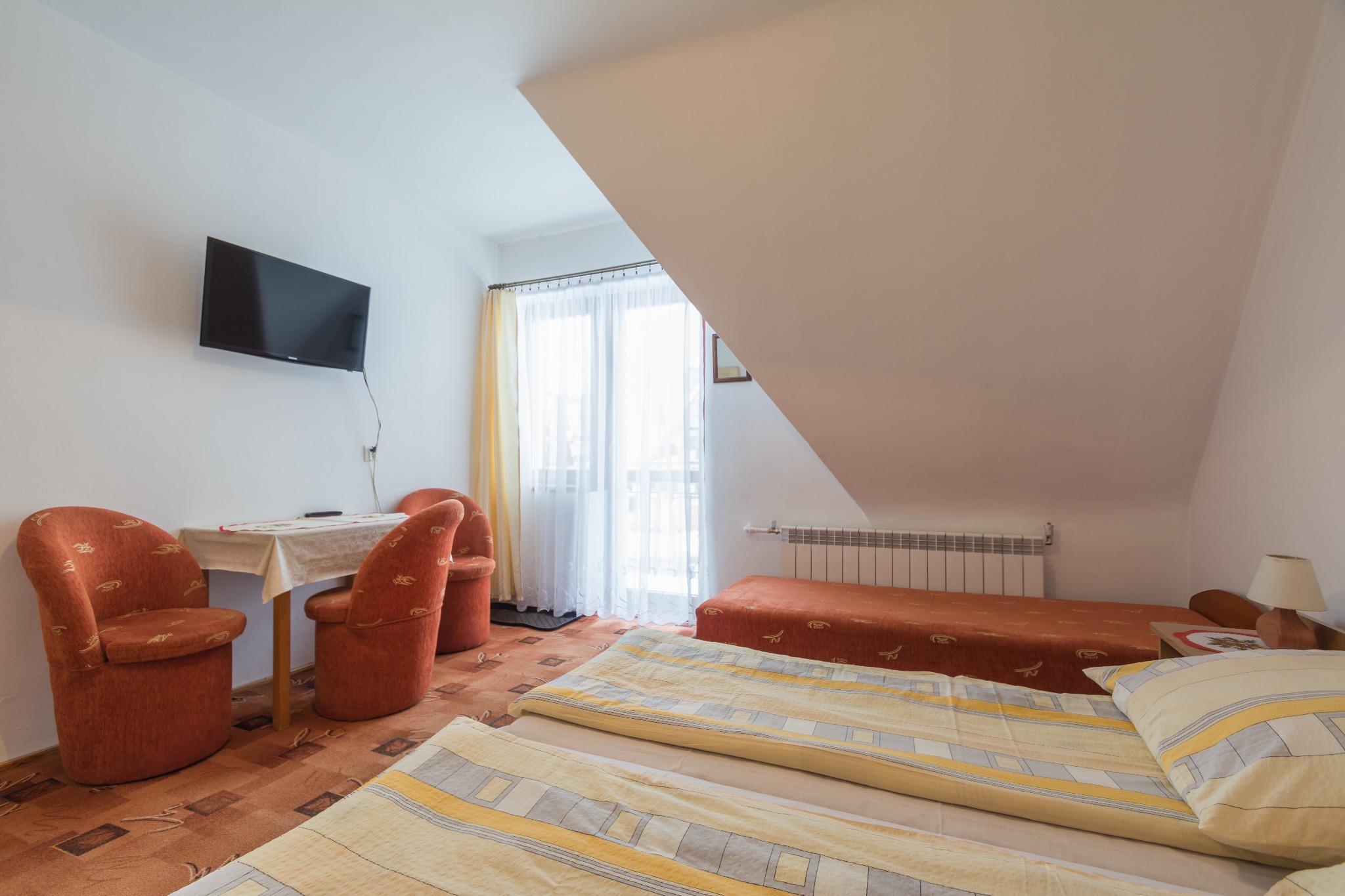 Jasne wnętrze pokoju na poddaszu z dwoma łóżkami, stolikiem i telewizorem, widoczne skosy sufitu i okno balkonowe.