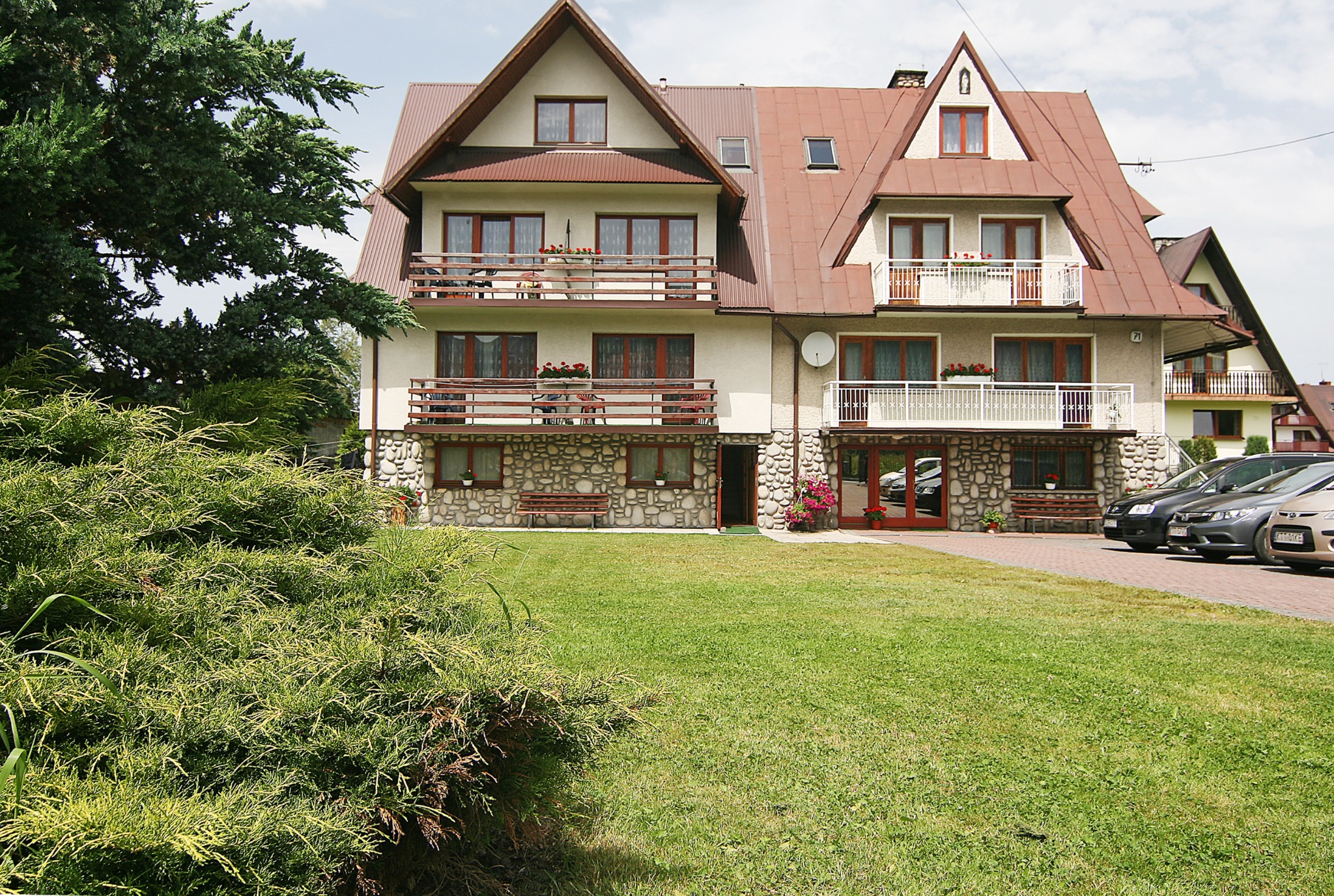 Dwupiętrowy budynek z beżową elewacją, brązowym dachem i balkonami z kwiatami, z kamiennym parterem i zadbanym trawnikiem przed budynkiem.
