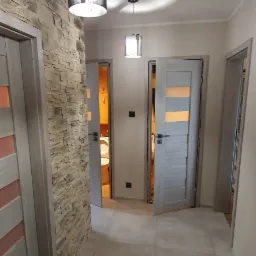 Korytarz po remoncie z kamienną ścianą, szarymi drzwiami z pomarańczowymi akcentami i dwiema lampami sufitowymi, z widokiem na otwartą łazienkę.