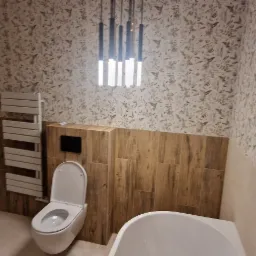 Nowoczesna łazienka z wiszącą toaletą, grzejnikiem drabinkowym i oświetleniem wiszącym, wykończona płytkami imitującymi drewno i tapetą w roślinny wzór.