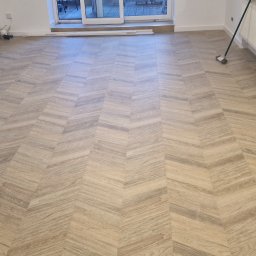Wol-Bud - Ułożona pod kątem podłoga drewnopodobna w jasnym, pustym pokoju z białymi ścianami i listwami przypodłogowymi, widoczne gniazdka elektryczne i grzejnik.