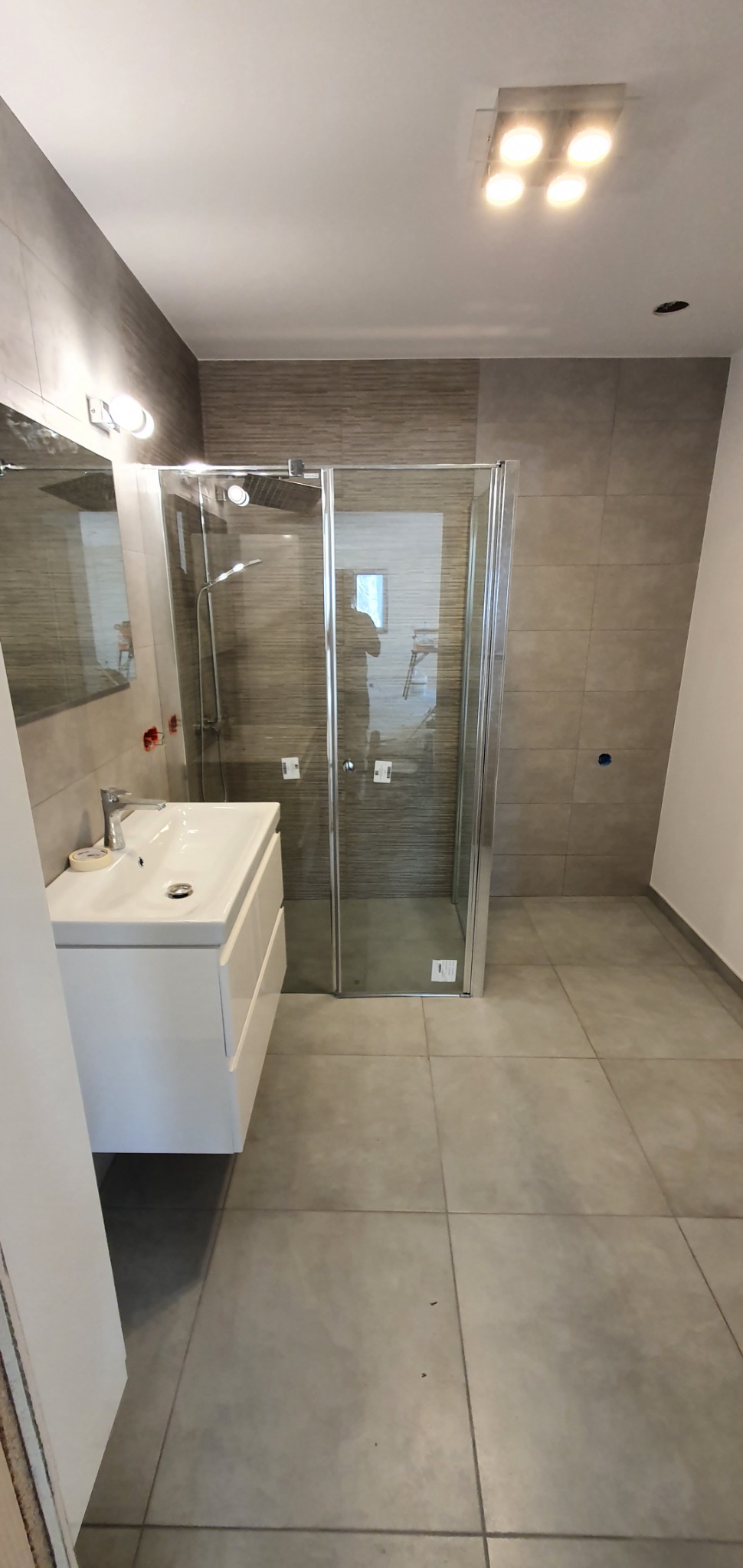 Nowoczesna łazienka po remoncie z kabiną prysznicową typu walk-in, umywalką z szafką w kolorze białym, szarą glazurą na ścianach i podłodze oraz oświetleniem sufitowym.