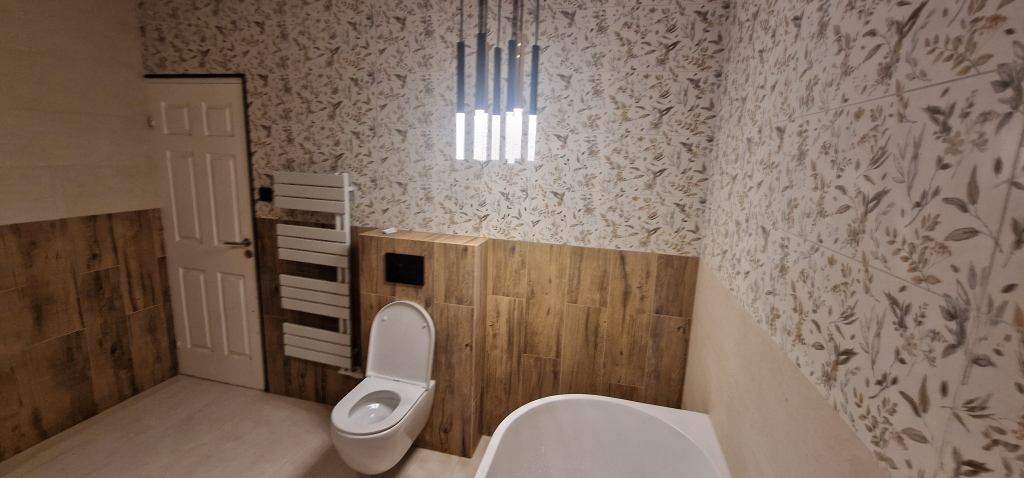 Nowoczesna łazienka z wiszącą toaletą, grzejnikiem drabinkowym i oświetleniem wiszącym, wykończona płytkami imitującymi drewno i tapetą w roślinny wzór.