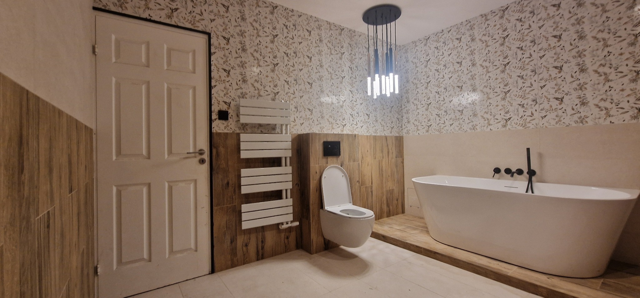 Nowoczesna łazienka z wolnostojącą białą wanną, wiszącą toaletą, grzejnikiem drabinkowym i wzorzystą tapetą w odcieniach beżu i brązu, oświetlona wiszącą lampą z podłużnymi kloszami.