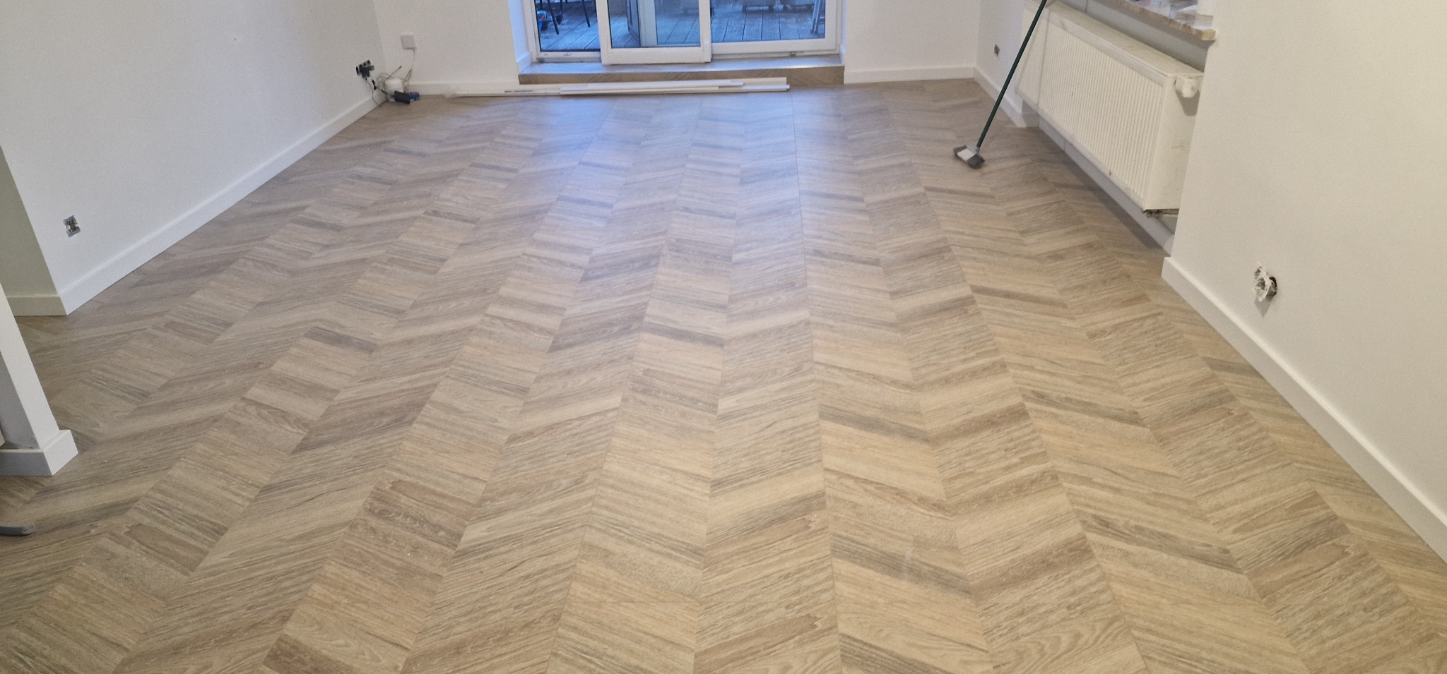 Ułożona pod kątem podłoga drewnopodobna w jasnym, pustym pokoju z białymi ścianami i listwami przypodłogowymi, widoczne gniazdka elektryczne i grzejnik.