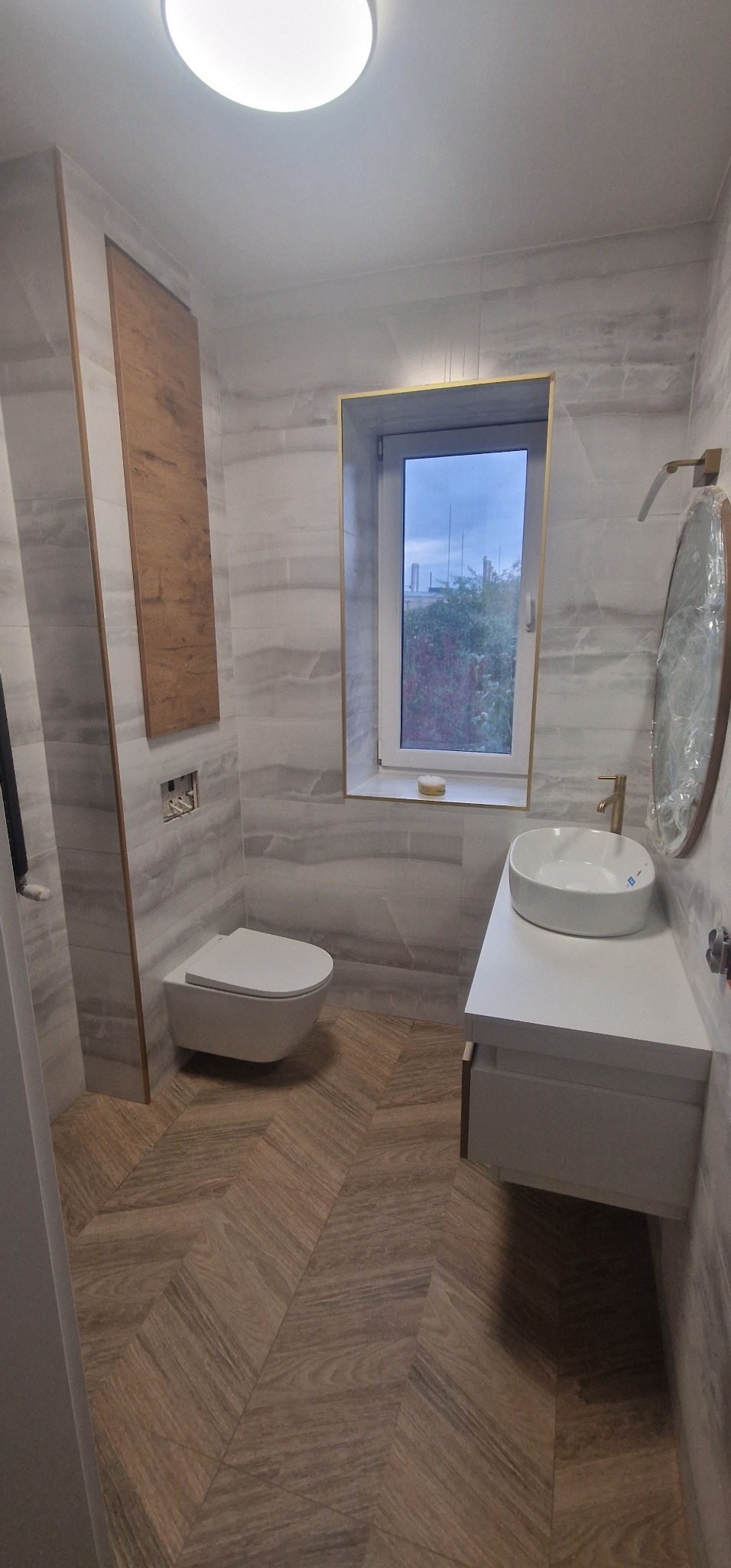 Nowoczesna łazienka z szarymi płytkami imitującymi marmur, drewnianą szafką, wiszącą toaletą i umywalką nablatową z baterią w kolorze złota, drewniana podłoga w jodełkę, okno z złotą ramą.