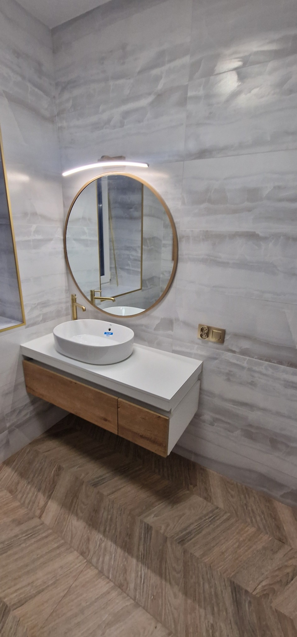 Nowoczesna łazienka z umywalką nablatową, okrągłym lustrem w złotej ramie, drewnianą szafką pod umywalką i podłogą ułożoną w jodełkę.