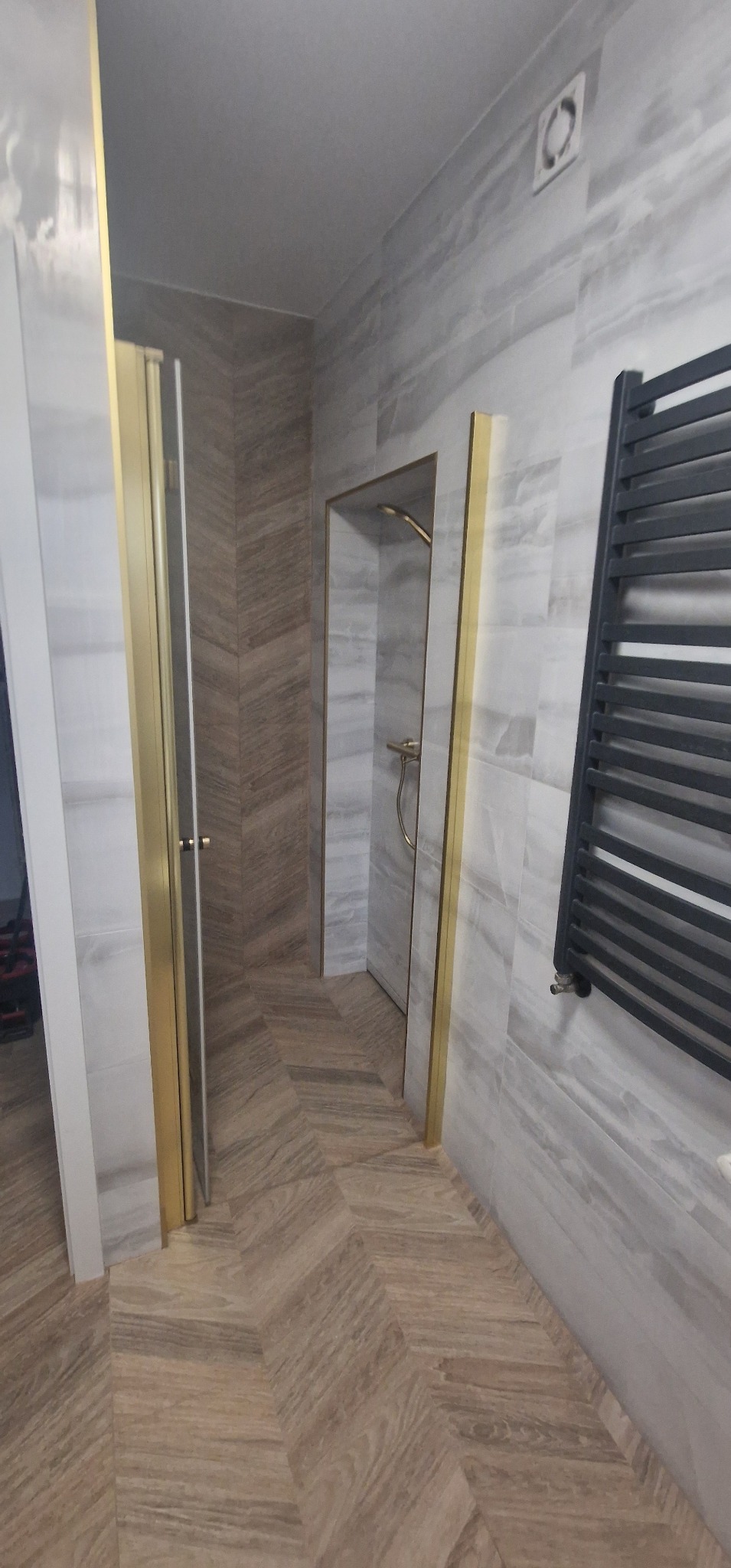 Nowoczesna łazienka z szarymi płytkami imitującymi marmur, podłoga ułożona w jodełkę, kabina prysznicowa z złotymi ramami i grzejnik drabinkowy.