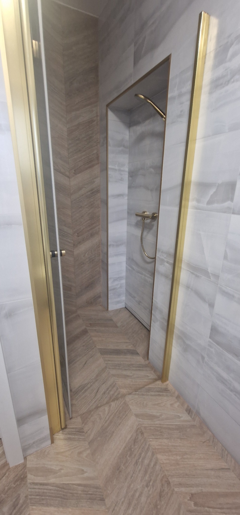 Wykończona łazienka z kabiną prysznicową typu walk-in, jasnymi płytkami imitującymi marmur na ścianach i podłodze ułożonej w jodełkę, z elementami wykończeniowymi w kolorze złotym.
