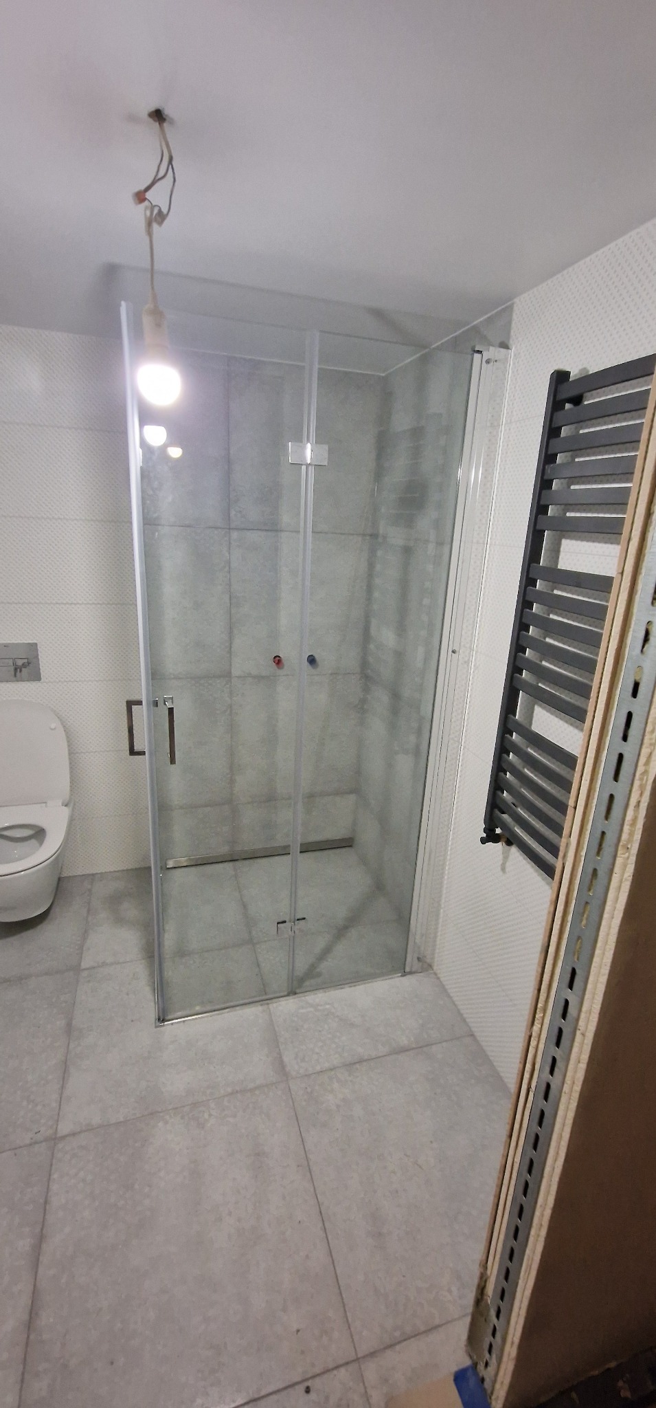 Szklana kabina prysznicowa z dwuskrzydłowymi drzwiami w wyremontowanej łazience z szarymi płytkami, widoczny grzejnik drabinkowy i toaleta, niedokończone oświetlenie sufitowe.