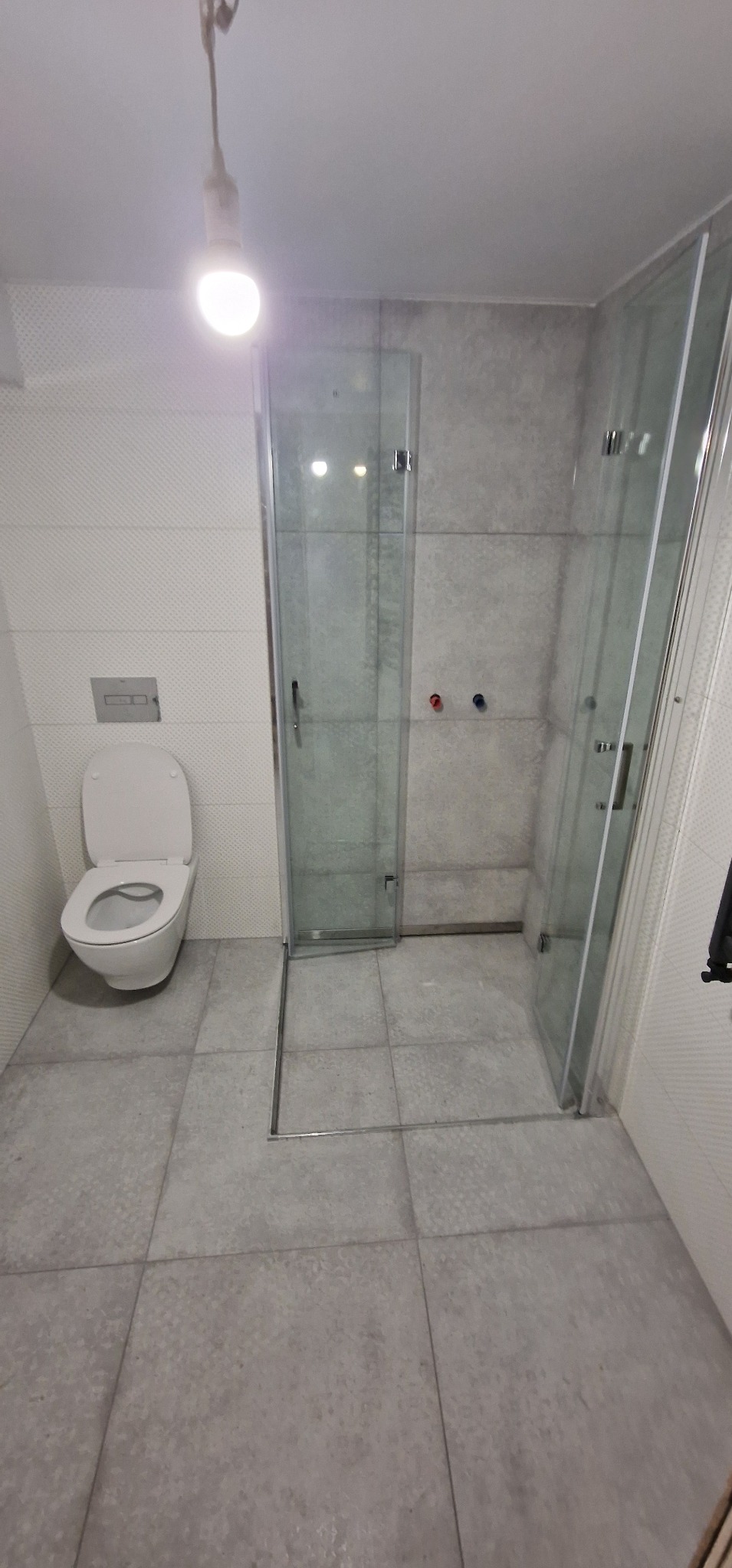 Nowoczesna łazienka z kabiną prysznicową typu walk-in, wiszącą toaletą i szarą płytką podłogową, oświetlona pojedynczą żarówką.