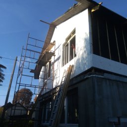 MAT-WOOD - Budowa domu: ściana zewnętrzna w trakcie ocieplania styropianem, widoczne rusztowanie i drabina oparta o ścianę, błękitne niebo w tle.