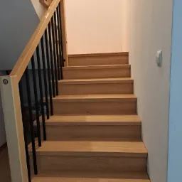 Drewniane schody z czarną balustradą w jasnym wnętrzu. Widok na stopnie i fragment ściany z wyłącznikiem.