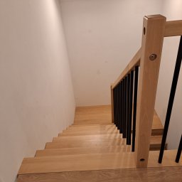 MAT-WOOD - Drewniane schody z czarną balustradą w jasnym wnętrzu. Widoczny fragment kartonu na podłodze, sugerujący etap wykończeniowy. Górne oświetlenie punktowe.