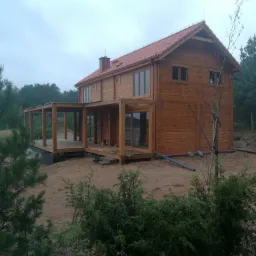 Dwukondygnacyjny dom z bali z odkrytym tarasem, brązowe drewno, czerwony dach. Wokół drzewa i krzewy, pochmurne niebo. Widok z zewnątrz, w trakcie budowy.