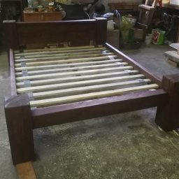 MAT-WOOD - Rama łóżka z ciemnego drewna z listwami, widoczna w warsztacie. Solidna konstrukcja, surowe wykończenie, detale połączeń stolarskich. Ujęcie z niskiej perspektywy.