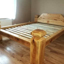 MAT-WOOD - Drewniane łóżko z surowego drewna, z wyraźnymi słojami i sękami, na parkiecie. Widoczne masywne nogi i zagłówek o geometrycznym kształcie. Naturalne oświetlenie.