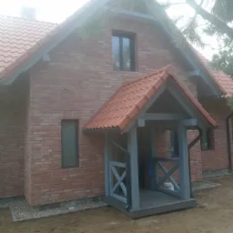 Dwukondygnacyjny dom z czerwonej cegły z zadaszonym wejściem w szarym kolorze i czerwoną dachówką, widok z zewnątrz na niezagospodarowany teren.