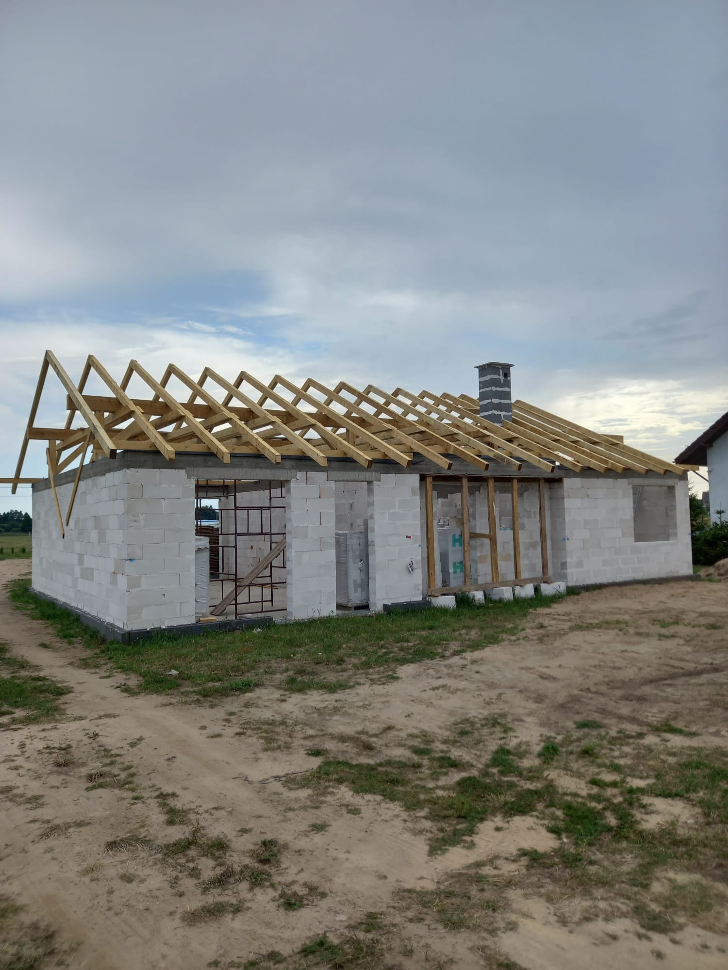 Niedokończony budynek w stanie surowym otwartym z widoczną konstrukcją dachu i kominem z szarej cegły, w tle pochmurne niebo.