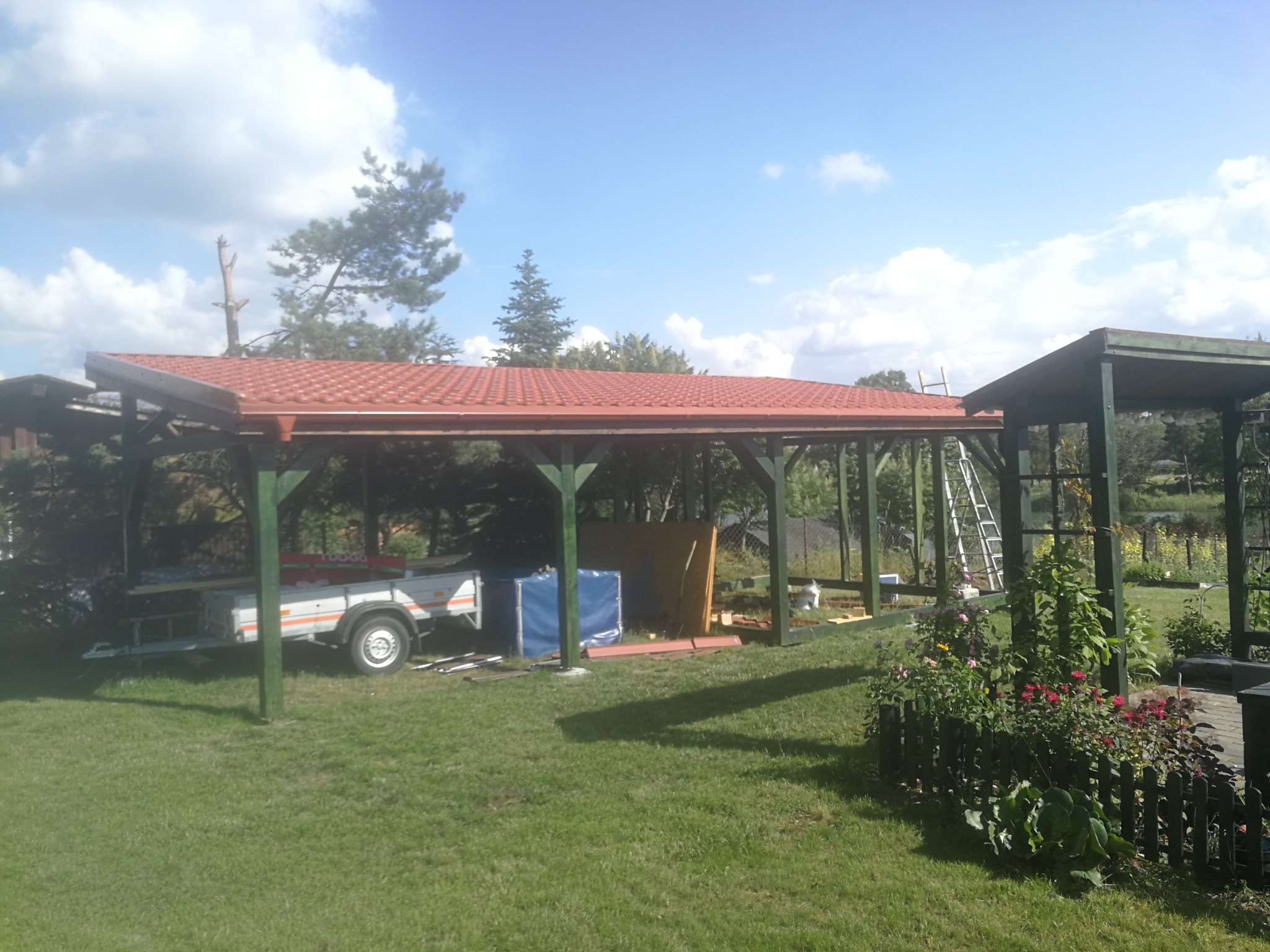 Montaż zadaszenia tarasu z czerwoną dachówką, widoczna przyczepka z materiałami budowlanymi i drabina oparta o konstrukcję.