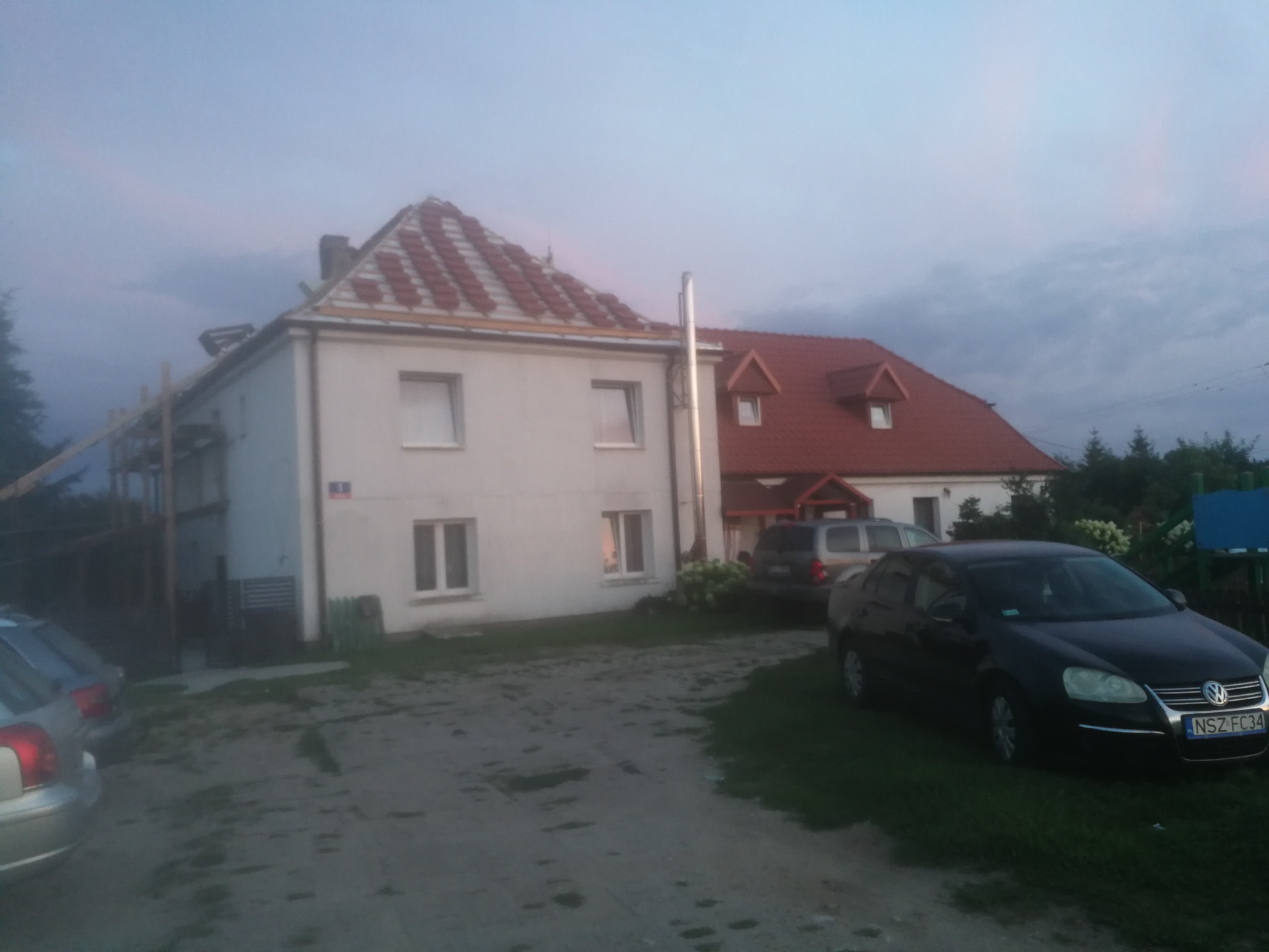 Dwupiętrowy dom z częściowo wymienionym pokryciem dachowym z czerwonej dachówki, widoczne rusztowania i zaparkowane samochody na posesji.