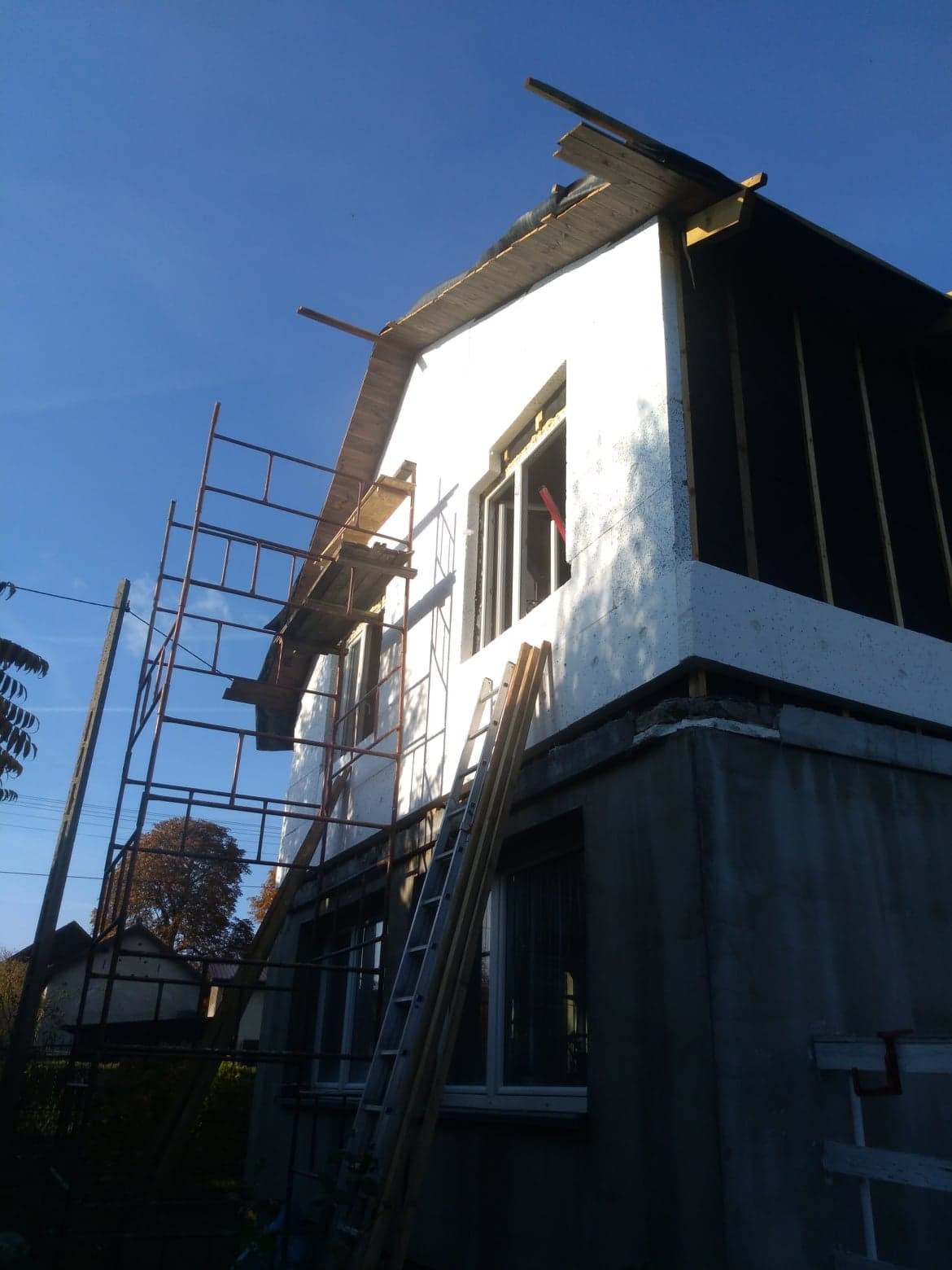 Budowa domu: ściana zewnętrzna w trakcie ocieplania styropianem, widoczne rusztowanie i drabina oparta o ścianę, błękitne niebo w tle.