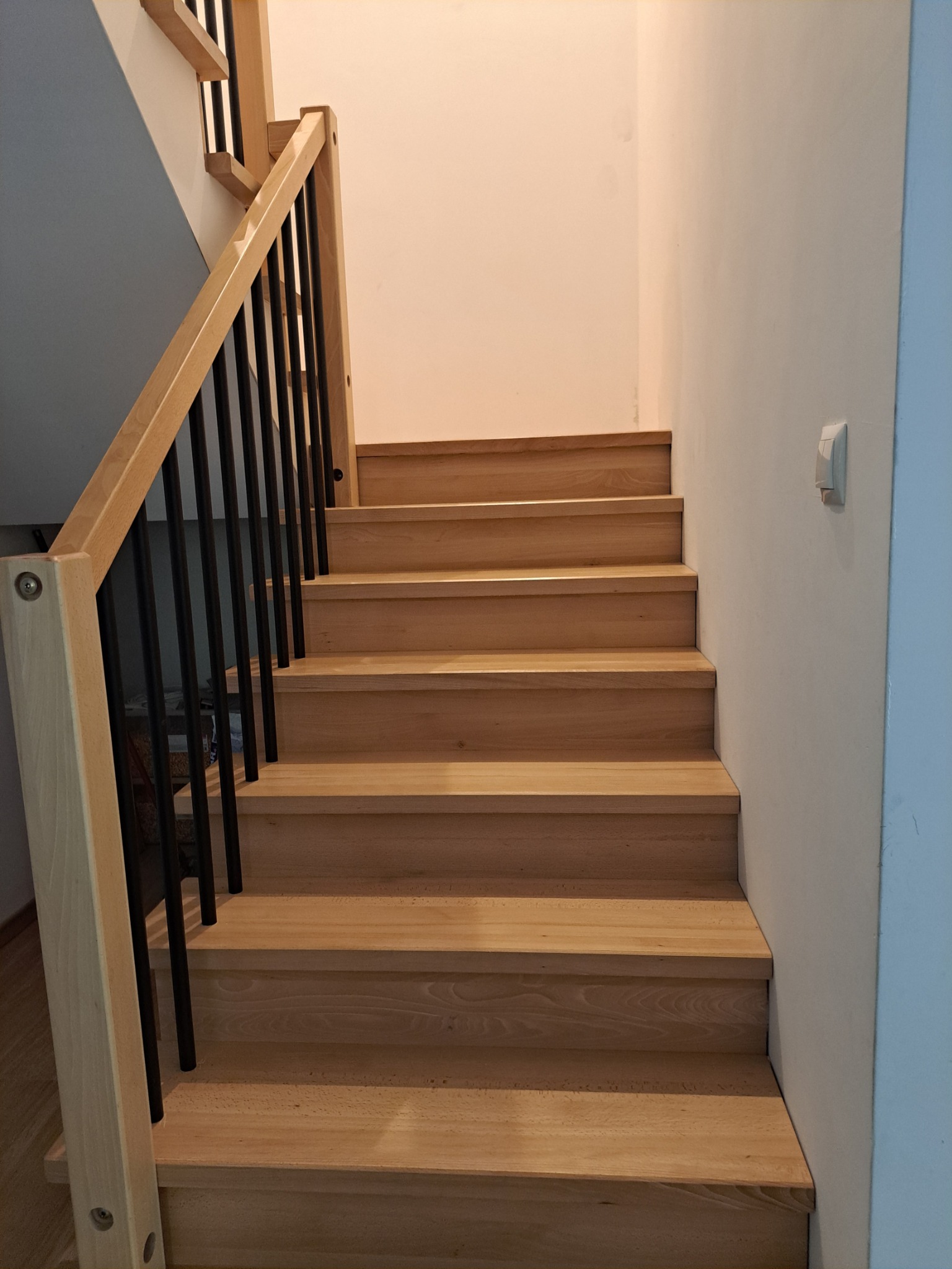 Drewniane schody z czarną balustradą w jasnym wnętrzu. Widok na stopnie i fragment ściany z wyłącznikiem.