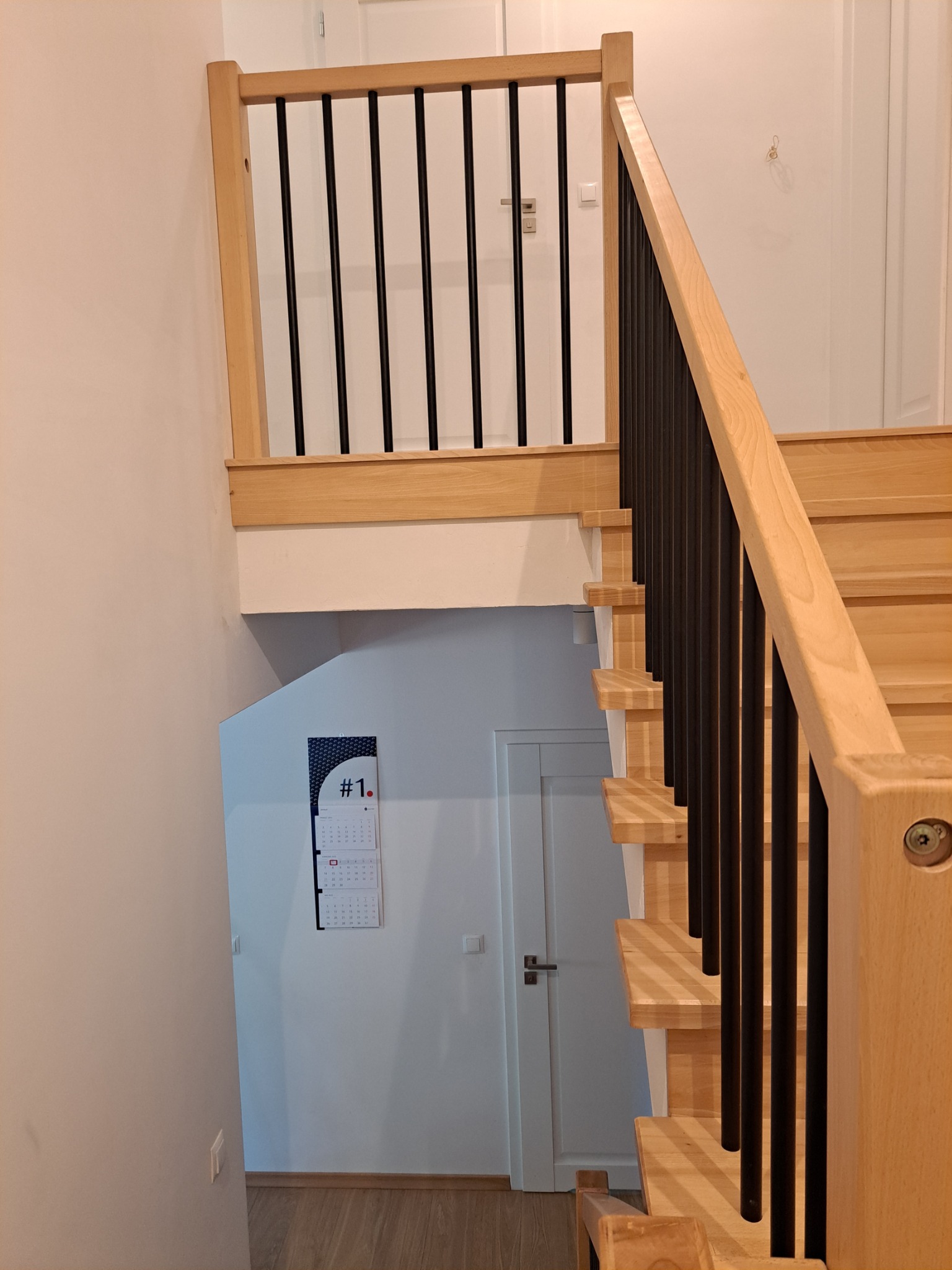 Jasne drewniane schody z czarnymi, metalowymi balustradami, widziane z góry. Na ścianie wisi kalendarz. Minimalistyczny design.