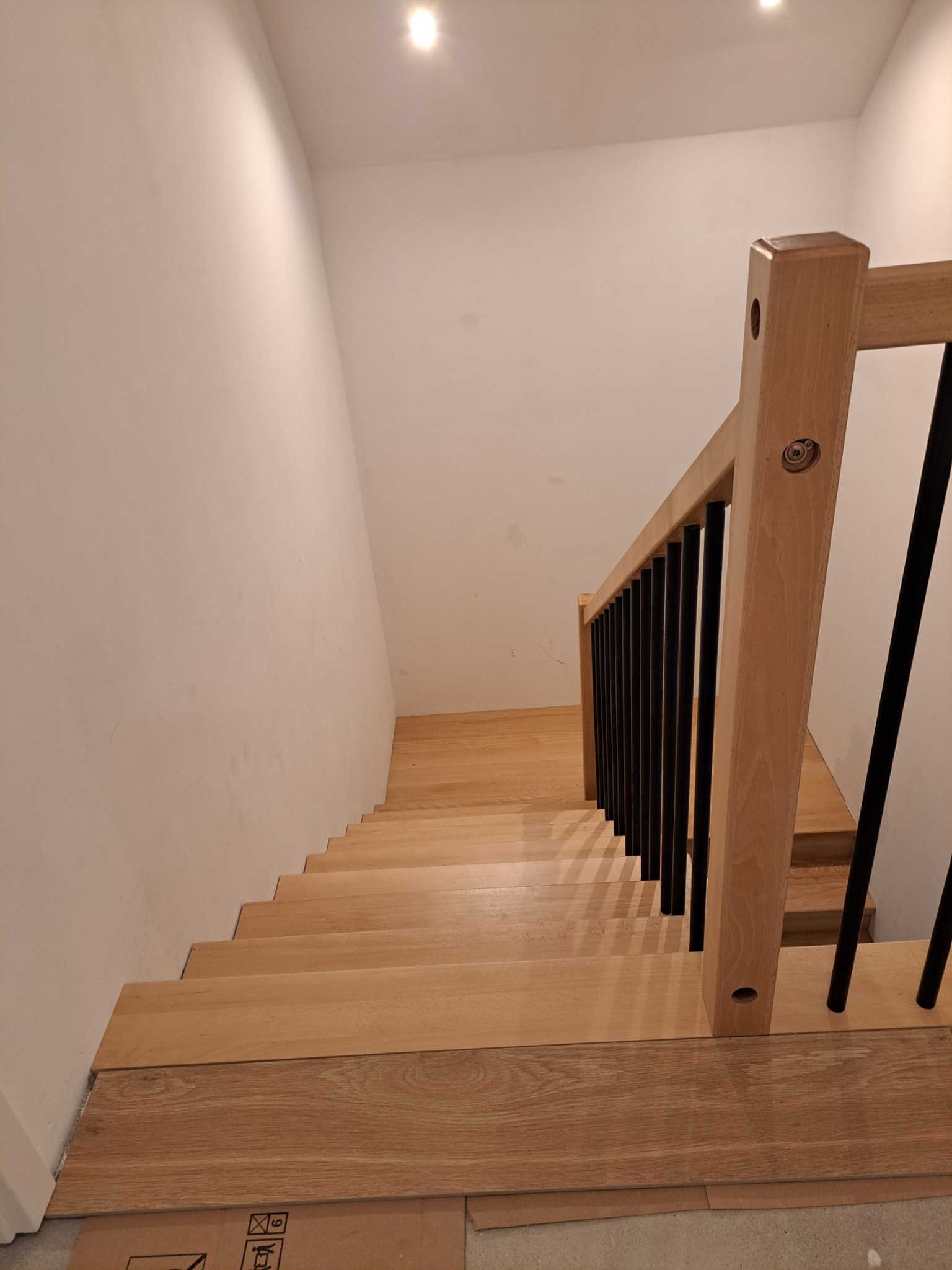 Drewniane schody z czarną balustradą w jasnym wnętrzu. Widoczny fragment kartonu na podłodze, sugerujący etap wykończeniowy. Górne oświetlenie punktowe.