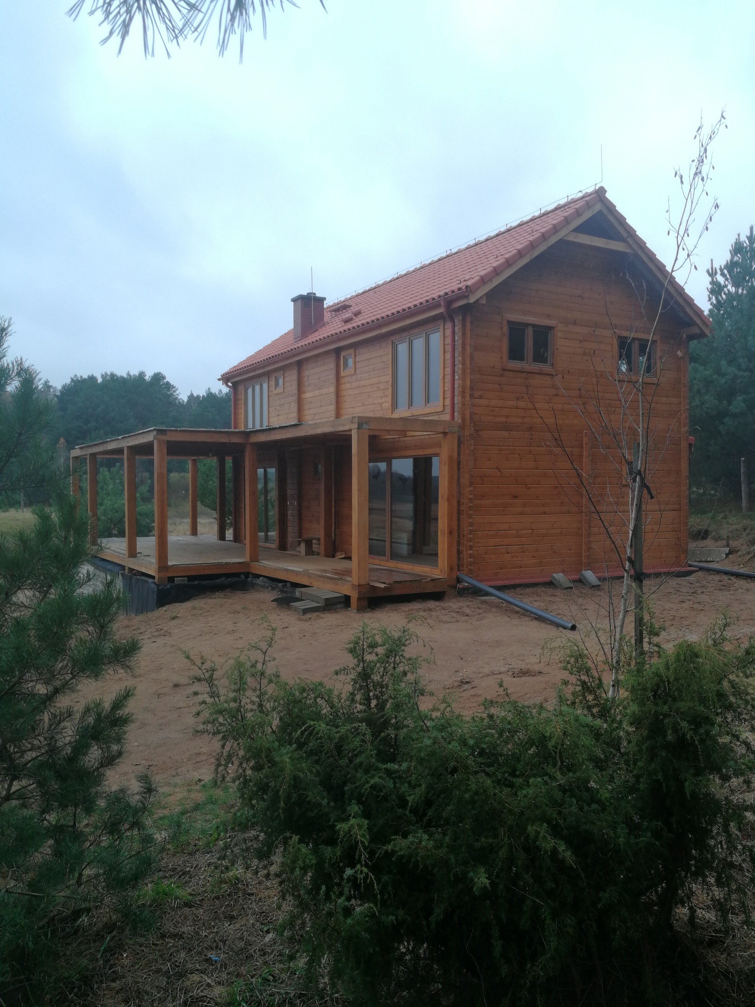 Dwukondygnacyjny dom z bali z odkrytym tarasem, brązowe drewno, czerwony dach. Wokół drzewa i krzewy, pochmurne niebo. Widok z zewnątrz, w trakcie budowy.