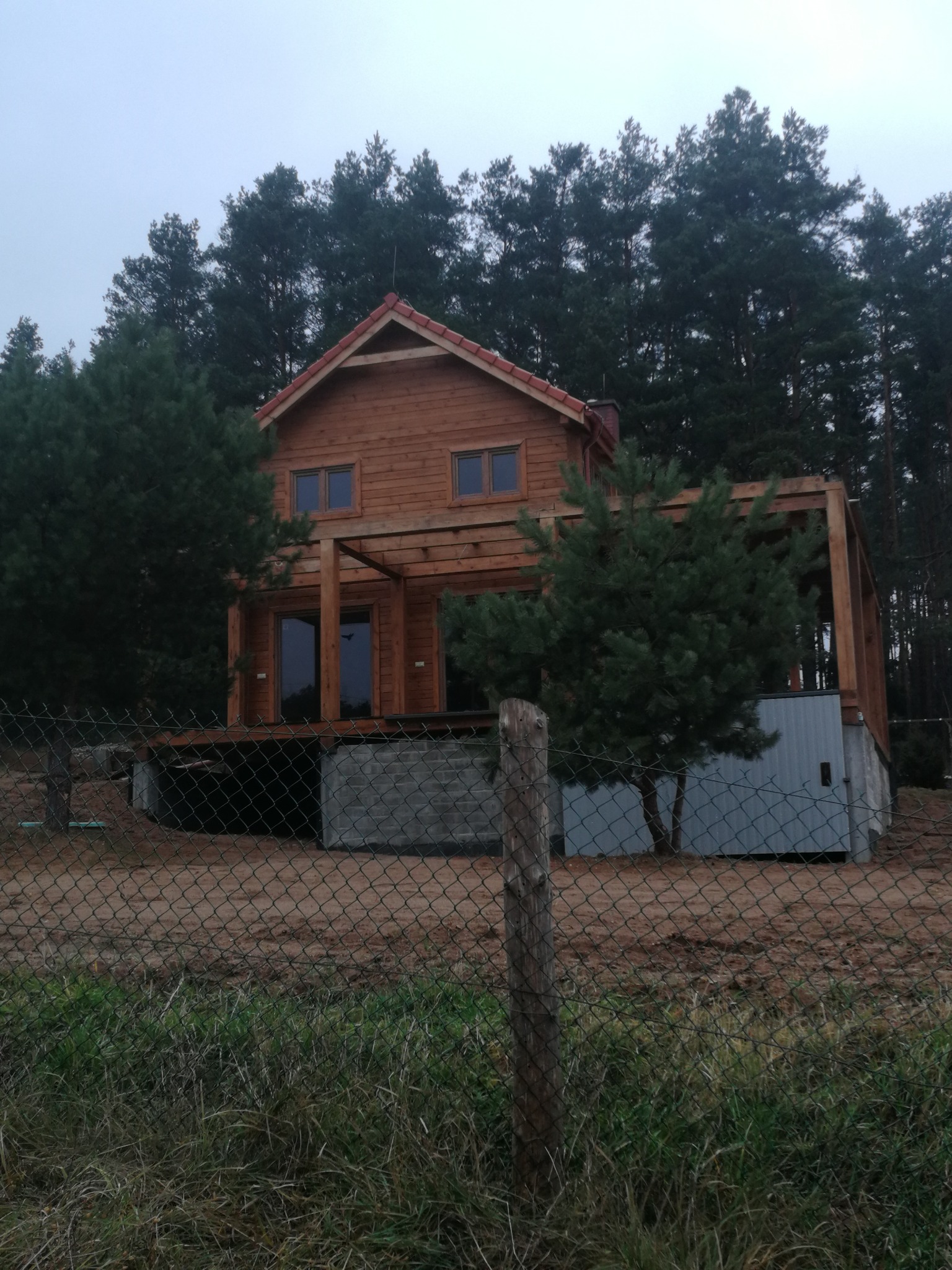 Drewniany dom szkieletowy z poddaszem i pergolą, otoczony lasem, widziany zza ogrodzenia. Dom na fundamencie z bloczków, z widoczną instalacją pod podłogą.