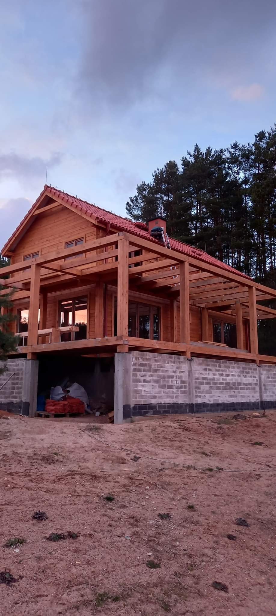 Surowy stan domu szkieletowego z czerwonym dachem, widoczna konstrukcja drewniana i fundament z bloczków. W tle las i niebo o zmierzchu.