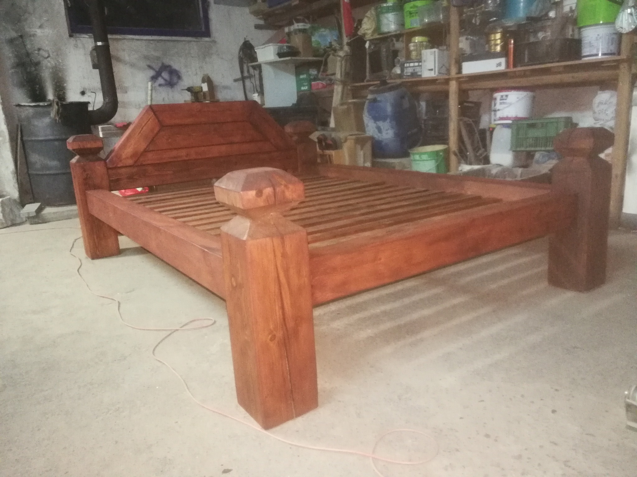 Drewniane łóżko w surowym wnętrzu warsztatu, z widoczną strukturą drewna i prostym zagłówkiem. W tle półki z narzędziami i farbami.