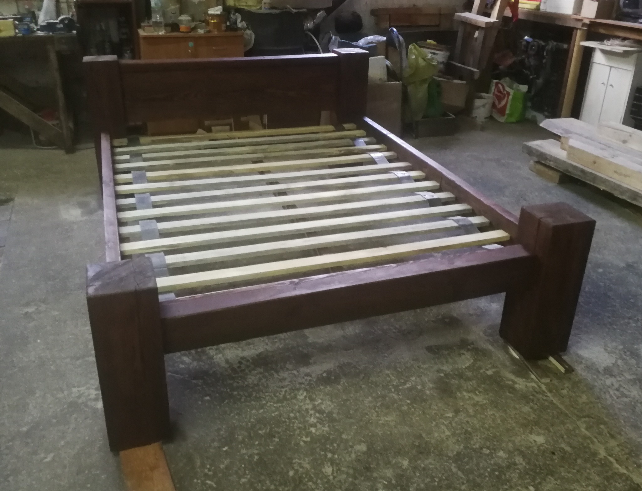 Rama łóżka z ciemnego drewna z listwami, widoczna w warsztacie. Solidna konstrukcja, surowe wykończenie, detale połączeń stolarskich. Ujęcie z niskiej perspektywy.