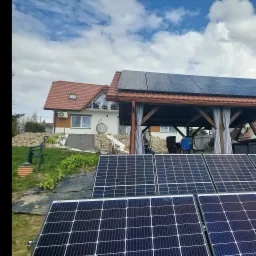 Panele słoneczne zamontowane na dachu altany w ogrodzie, w tle dom jednorodzinny z klimatyzatorem na ścianie.