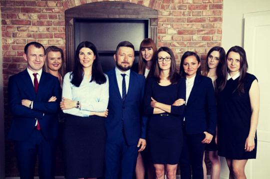 Grupa prawników, mężczyźni w garniturach z krawatami, kobiety w sukienkach i marynarkach, stojąca na tle ściany z cegły i ciemnych drzwi z ozdobnym obramowaniem.