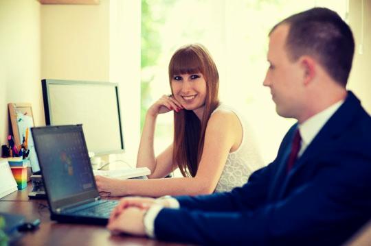 Kobieta z uśmiechem i mężczyzna w garniturze siedzą przy biurku z laptopem i monitorem; naturalne światło z okna w tle.