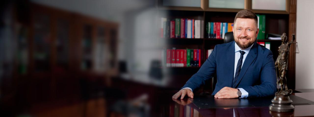 Uśmiechnięty adwokat w niebieskim garniturze siedzi za biurkiem w gabinecie, obok statuetka Temidy i regał z książkami.