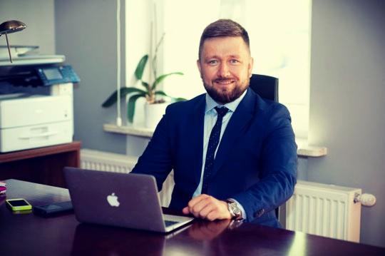 Uśmiechnięty mężczyzna w niebieskim garniturze siedzi za biurkiem z laptopem Apple, telefonem i zegarkiem na nadgarstku, w tle biuro z rośliną w doniczce i drukarką.