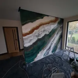 Ściana w trakcie dekoracyjnego malowania z efektem marmuru w odcieniach zieleni, bieli i brązu, zabezpieczona folią podłoga, widoczne narzędzia malarskie i drabina.