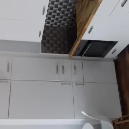 Nowoczesna kuchnia z białymi szafkami, drewnianym blatem i geometrycznym wzorem na ścianie nad blatem, widoczny fragment drewnianej podłogi i białego krzesła.