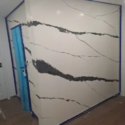 Ściana w trakcie dekoracyjnego wykończenia imitującego marmur z widocznymi smugami farby, zabezpieczona niebieską taśmą malarską przy suficie, podłodze i futrynie drzwiowej z folią ochronną.