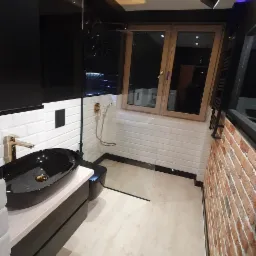 Nowoczesna łazienka z czarną umywalką nablatową, złotą armaturą, białymi płytkami metra i ścianą z cegły, oświetlona designerską lampą sufitową.