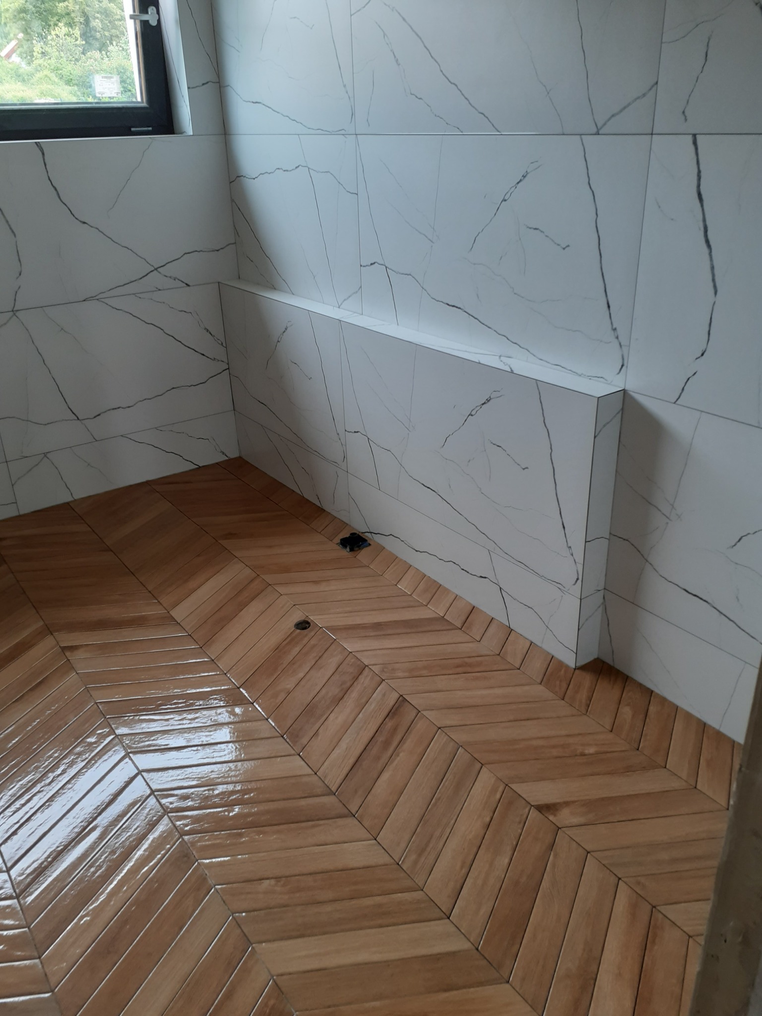 Ukończona łazienka w Redzie z drewnopodobną podłogą ułożoną w jodełkę i białymi płytkami imitującymi marmur na ścianach, widoczna półka w ścianie.