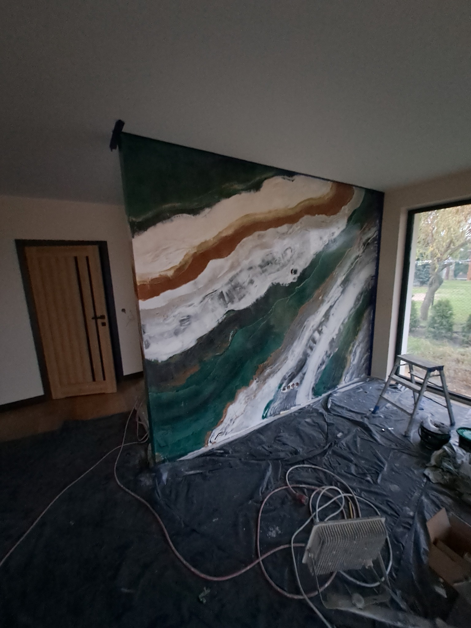 Ściana w trakcie dekoracyjnego malowania z efektem marmuru w odcieniach zieleni, bieli i brązu, zabezpieczona folią podłoga, widoczne narzędzia malarskie i drabina.