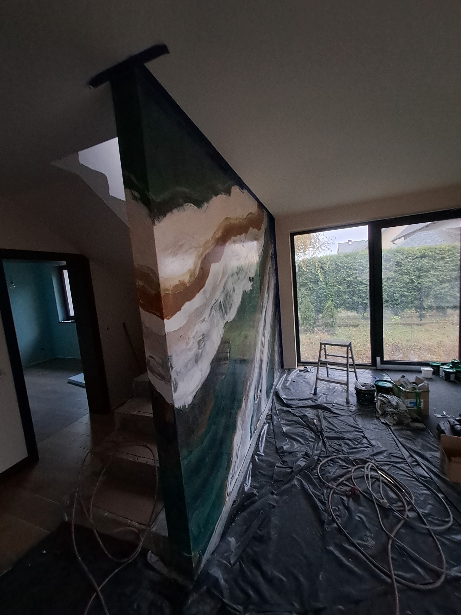 Ściana w trakcie dekoracyjnego malowania z efektem marmuru, zabezpieczona folią podłoga, drabina i puszki z farbami w pomieszczeniu z oknem na ogród.