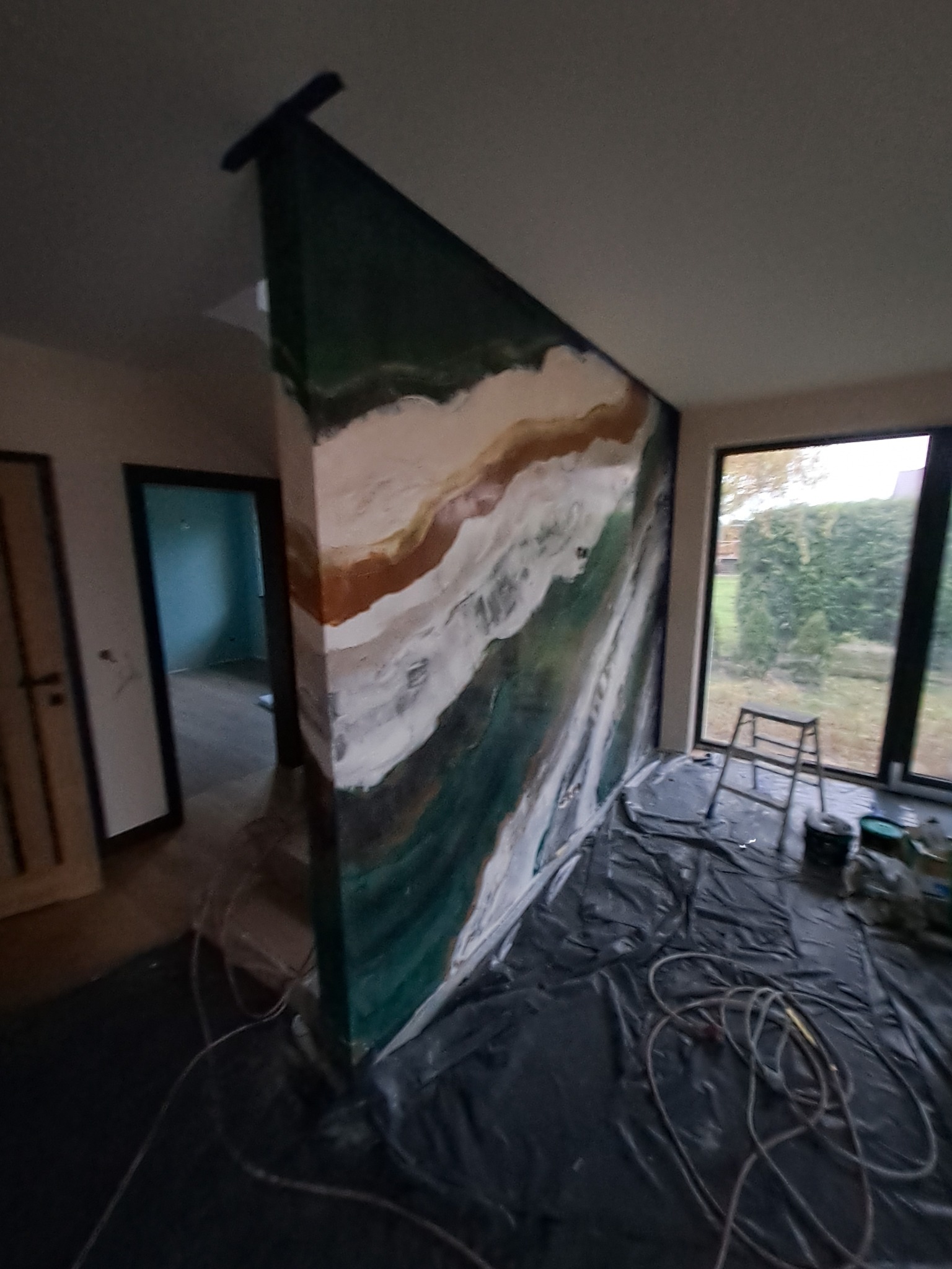 Ściana działowa z dekoracyjnym malowaniem imitującym przekrój geologiczny, zabezpieczona folią malarską, widoczna drabina i okno.