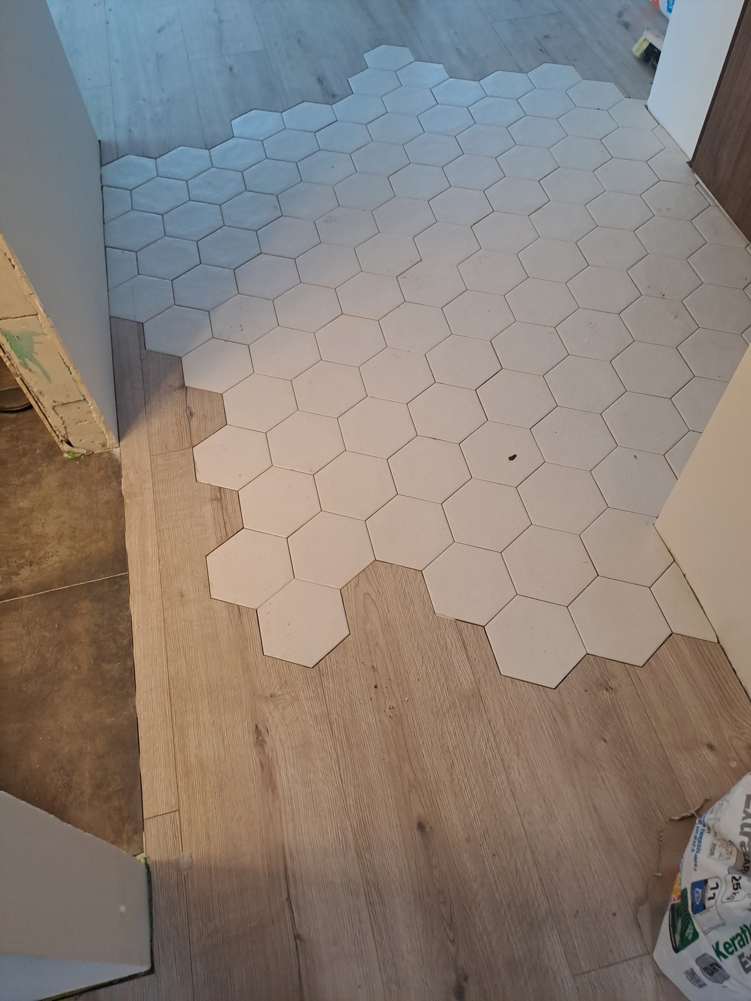 Układanie białych, heksagonalnych płytek na podłodze, częściowo pokrywających drewnopodobne panele. Widoczny fragment worka z zaprawą klejową Kerakoll.