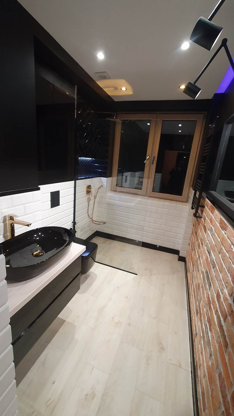 Nowoczesna łazienka z czarną umywalką nablatową, złotą armaturą, białymi płytkami metra i ścianą z cegły, oświetlona designerską lampą sufitową.