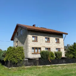 Dwupiętrowy dom z szarego pustaka z brązowym dachem i rynnami, widoczny z poziomu gruntu w słoneczny dzień. Na pierwszym planie czarny płot i zieleń, po lewej stronie biały samochód dostawczy.